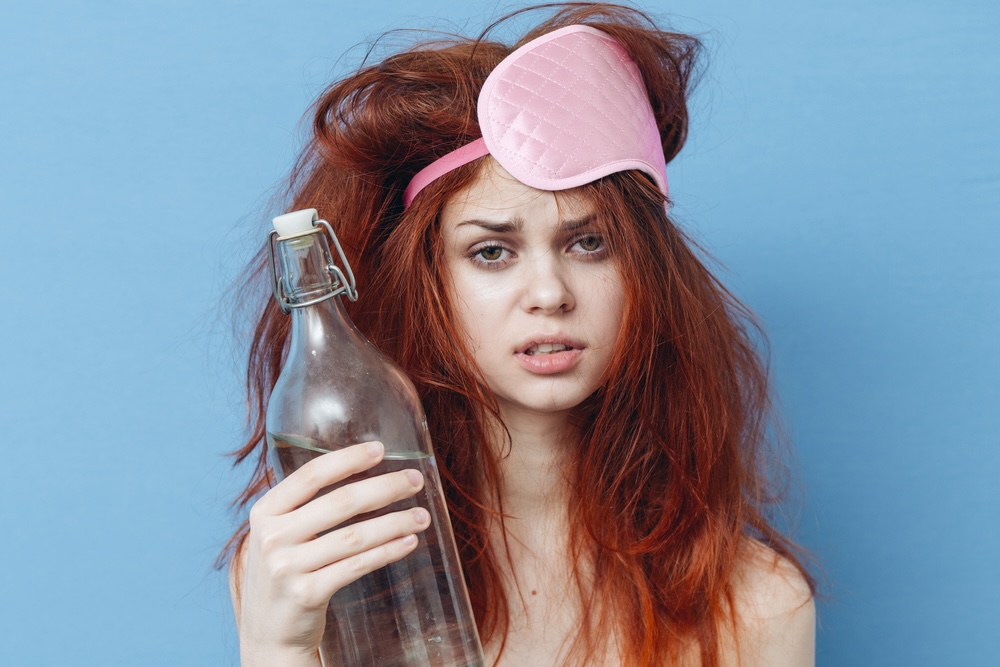 A woman with a hangover with red hair and pink sleeping mask holding a bottle of water