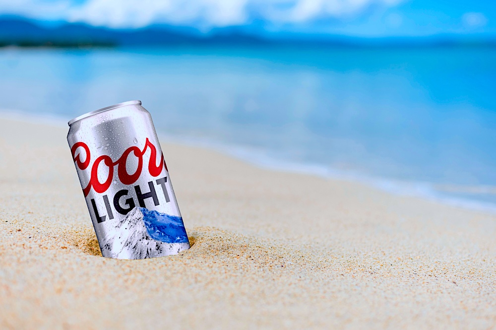 A can of Coors Light beer half-buried in white sand by the sea