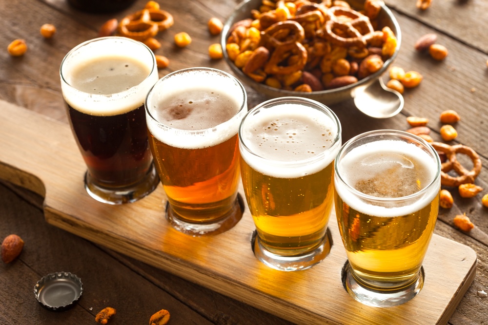 Four different types of beer with varying colors on a wooden tray and scatteredsnacks in the background