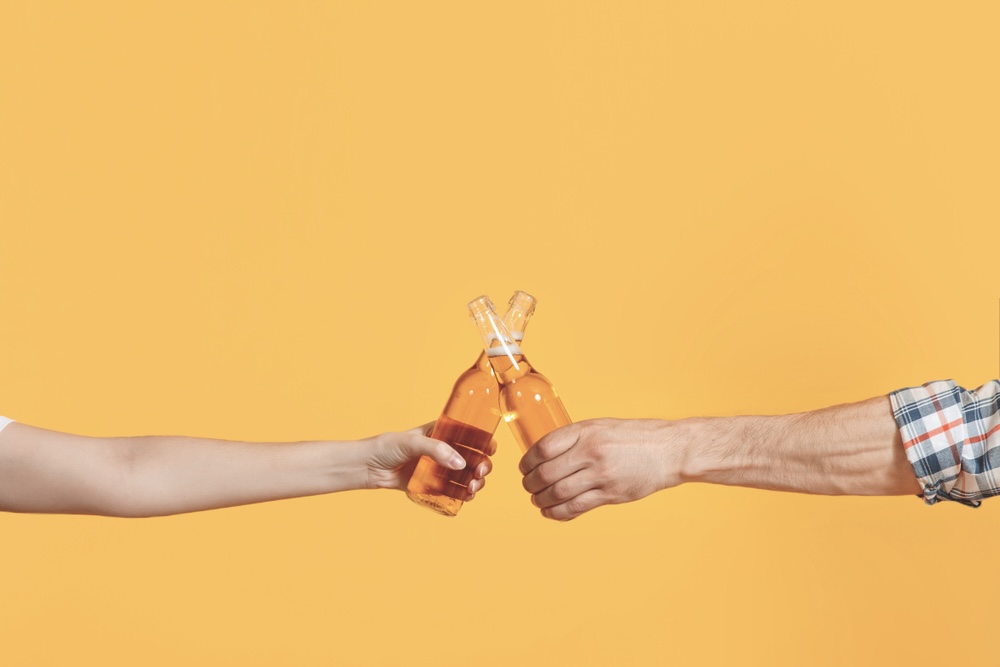 Two hands holding beers and clinking the bottles together on a yellow background