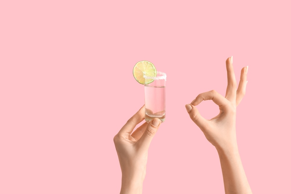 Female hands holding shot of tequila and showing OK gesture on pink background